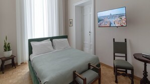 Ein Hotelzimmer mit Bett, Sessel, kleinem Tisch, Topfpflanze und Fernseher an der Wand.