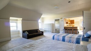 Una camera da letto con due letti, un divano scuro, una zona cucina visibile attraverso una porta aperta e un'ampia finestra che fa entrare luce naturale.