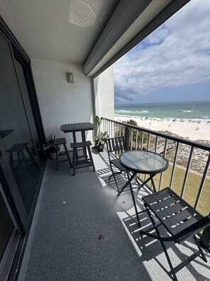 Uma varanda com mesa e cadeiras de vidro, com vista para uma praia com guarda-sóis e um céu nublado.