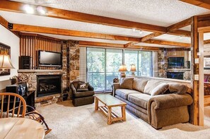 Ein gemütliches Wohnzimmer mit Kamin aus Stein, Holzbalken, einem großen Fenster und einer bequemen Couch.