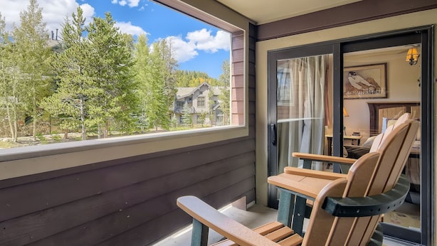 En balkon med træstole, en glasdør ind til et rum med en seng og en lampe, samt udsigt til træer og et hus.