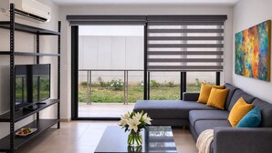 Een moderne woonkamer met een grijze bank, een zwarte salontafel, een televisie op een zwarte plank, en een groot raam met zonwering.