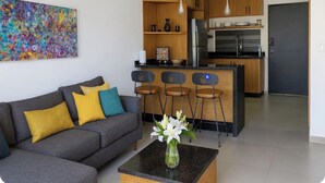 Een moderne woonkamer met een grijze bank, kleurrijke kussens, een salontafel met een bloevas, en een keuken met houten kastjes, een koelkast en barkrukken.
