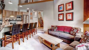 Eine moderne Küche mit Holzschränken, Edelstahlgeräten und einem Essbereich mit Holzsesseln. Eine Treppe führt zu einer oberen Ebene, und es gibt einen Wohnbereich mit einer Couch und Kissen.