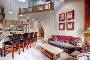 A modern kitchen with wooden cabinets, stainless steel appliances, and a dining area with wooden chairs. There is a staircase leading to an upper level, and a living area with a sofa and cushions. - Powder Ridge 208 - close to lifts and town! (Breckenridge)