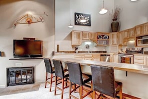A modern kitchen with wooden cabinets, a large island with bar stools, a flat-screen TV, and decorative wall art. - Powder Ridge 208 - close to lifts and town! (Breckenridge)