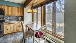 A kitchen with wooden cabinets, a stone backsplash, a dining table with chairs, and large windows offering a view of trees and a road. - Four O'Clock Lodge Condo- close to lifts and town! (Breckenridge)