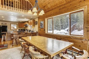 Un intérieur de cabine spacieux, avec une grande table à manger, des chaises en bois, un coin cuisine et de grandes fenêtres offrant une vue sur une forêt enneigée.