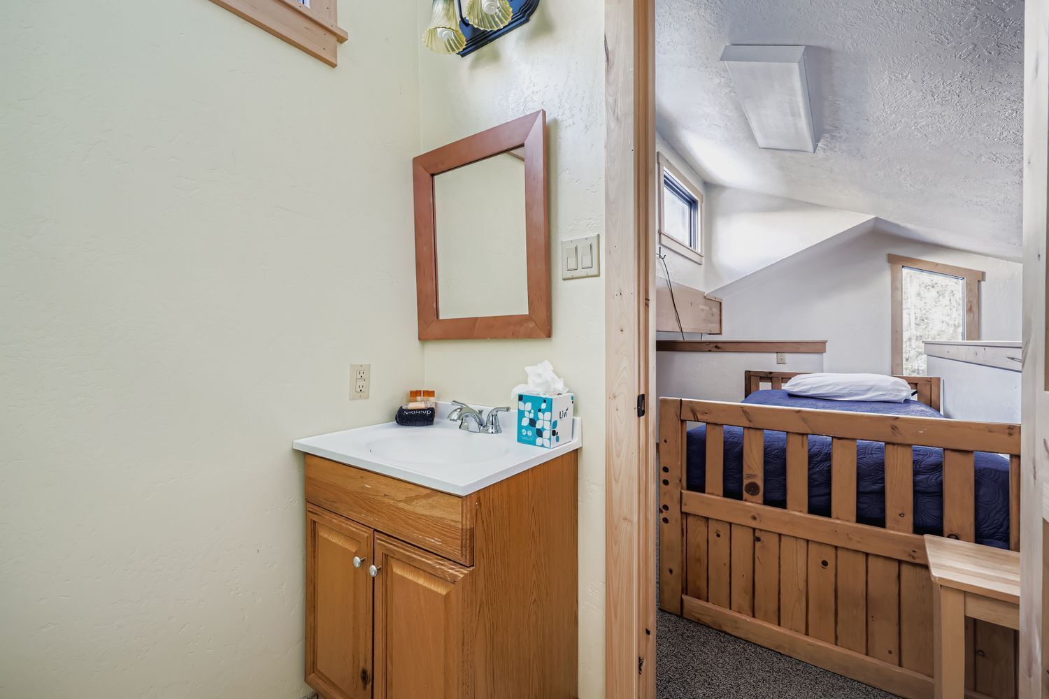 A compact room with a wooden bunk bed, a small bathroom with a sink and mirror, and a window providing natural light.