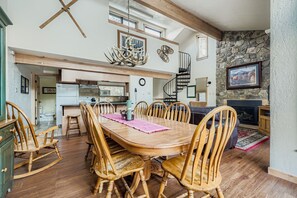Un vaste espace à manger avec une table et des chaises en bois, une cuisine à l’arrière-plan, une cheminée en pierre et un escalier menant à un niveau supérieur.