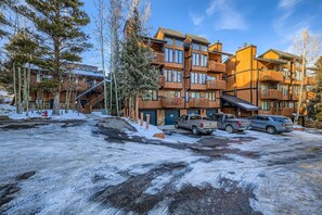 Un immeuble en bois à plusieurs étages, doté de balcons, une aire de stationnement avec plusieurs véhicules et un sol enneigé.