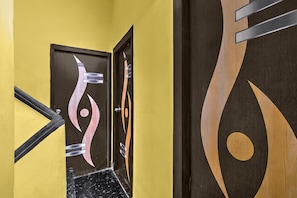 A hallway with a yellow wall, a black and white abstract door design, and a wooden door with a circular pattern. - Classic The Avenue Inn (Bidhannagar)