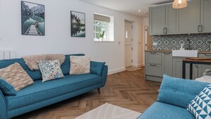 A modern living room with a blue sofa, a kitchen area with patterned backsplash, and two framed landscape paintings on the wall. - Shamrock (Exeter)