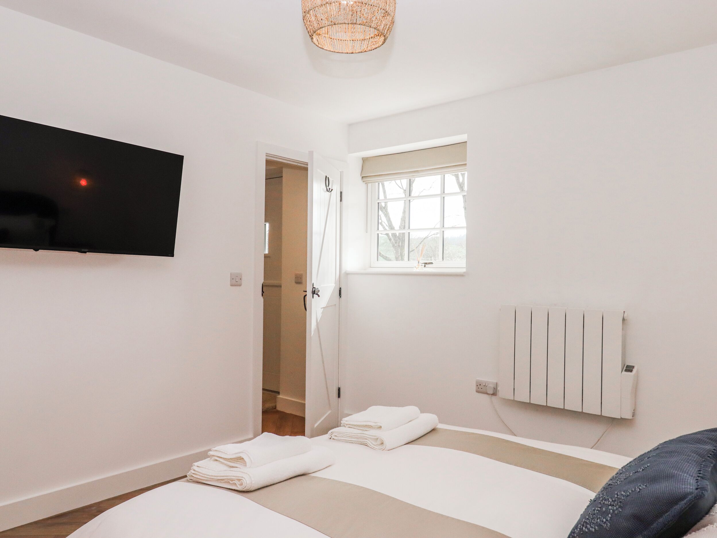 A modern bedroom with a flat-screen TV, a window with blinds, a radiator, and neatly folded towels on the bed.
