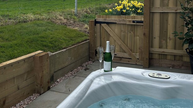 A hot tub with a bottle and glasses, surrounded by a wooden fence and a grassy area. - Shamrock (Exeter)