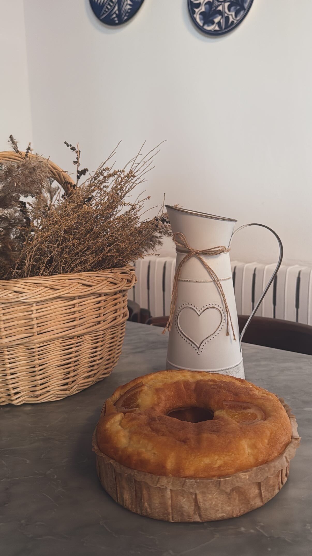 A wicker basket filled with dried plants, a white vase with a heart design, and a round wooden object on a table.