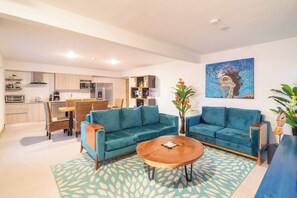 A modern living room with a blue sofa, a wooden coffee table, and a dining area with wicker chairs. - Sun, Style & Location | Viva Jacó Condo (Jacó)