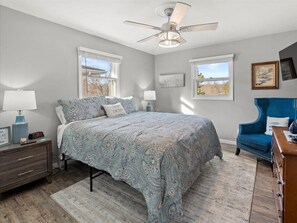 Ein Schlafzimmer mit einem großen Bett, einem Deckenventilator, einem Schminktisch, einem Stuhl und zwei Fenstern.
