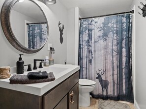 Ein Badezimmer mit weißem Waschbecken, dunklem Schrank und Spiegel. An der Wand ist eine Hirschsilhouette zu sehen, und ein Duschvorhang mit Waldmotiv.