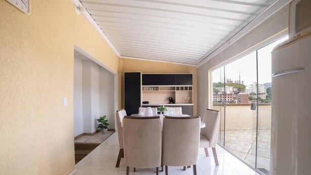 A dining area with a table and chairs, a kitchen area with shelves, and a view of buildings through large windows. - Apartments in Jd. Centenario in Pocos de Caldas - Clds (Poços de Caldas)