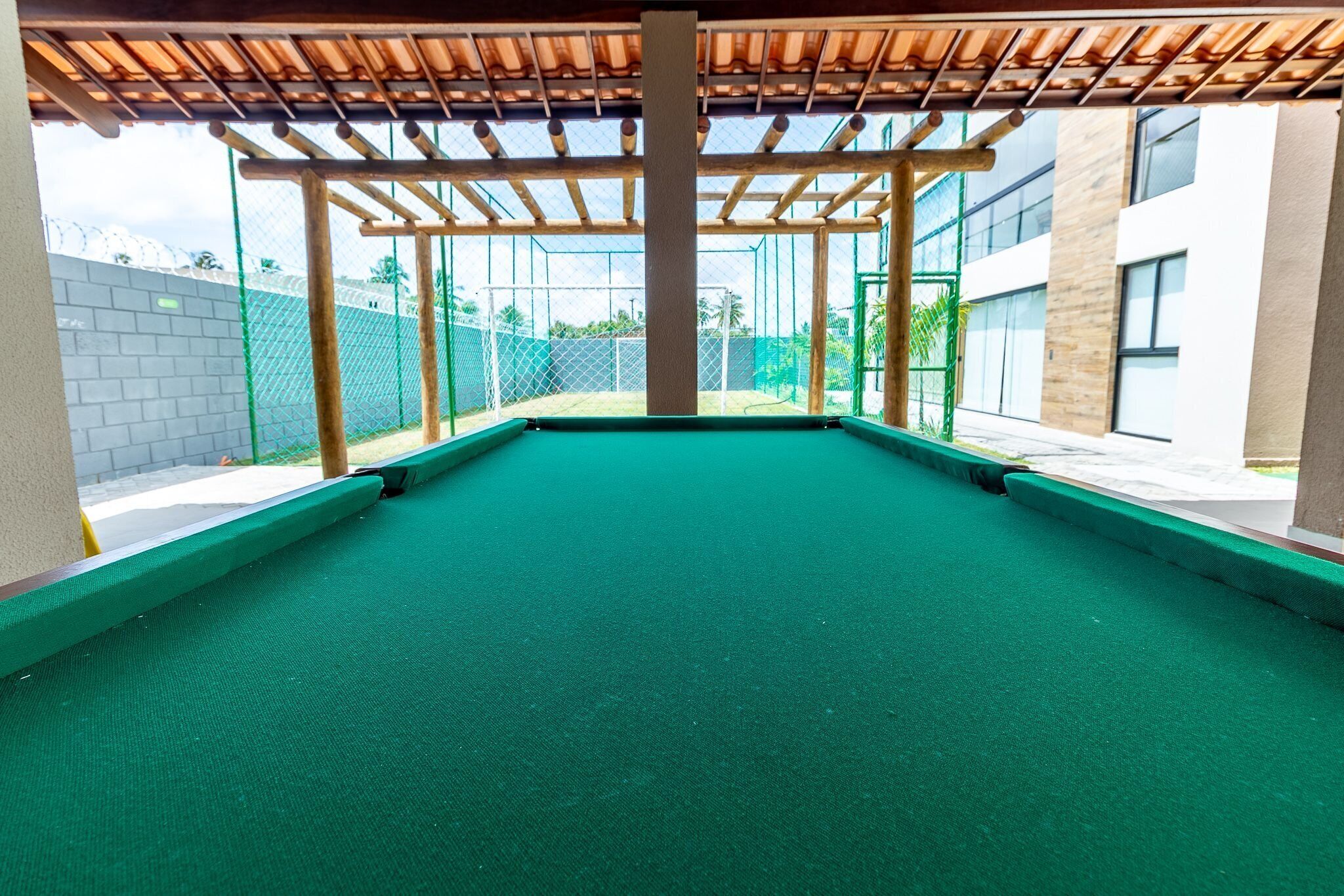 An indoor pool area with a green pool surface, wooden pergola overhead, and a view of a building with large windows and a brick wall in the background.