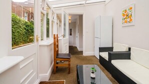 Ein sonnendurchflutetes Zimmer mit Glastür zu einem Garten, ein Holzstuhl, ein schwarzer Tisch mit Pflanze und ein weißes Sofa.