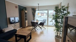 Una sala moderna con sofá, mesa de centro y comedor con sillas. Hay un televisor montado en la pared, una planta en maceta y un ventanal con vistas al exterior.