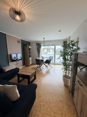 A modern living room with a sofa, coffee table, and a dining area with chairs. There is a TV mounted on the wall, a potted plant, and a large window with a view of the outside. - House Poseidon, vacation apartment POS01 (Cuxhaven)