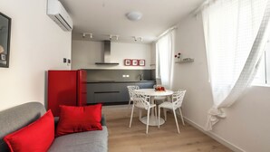En kompakt bolig med et tekøkken, et spiseområde med et rundt bord og stole, en grå sofa med røde puder og et vindue med hvide gardiner.