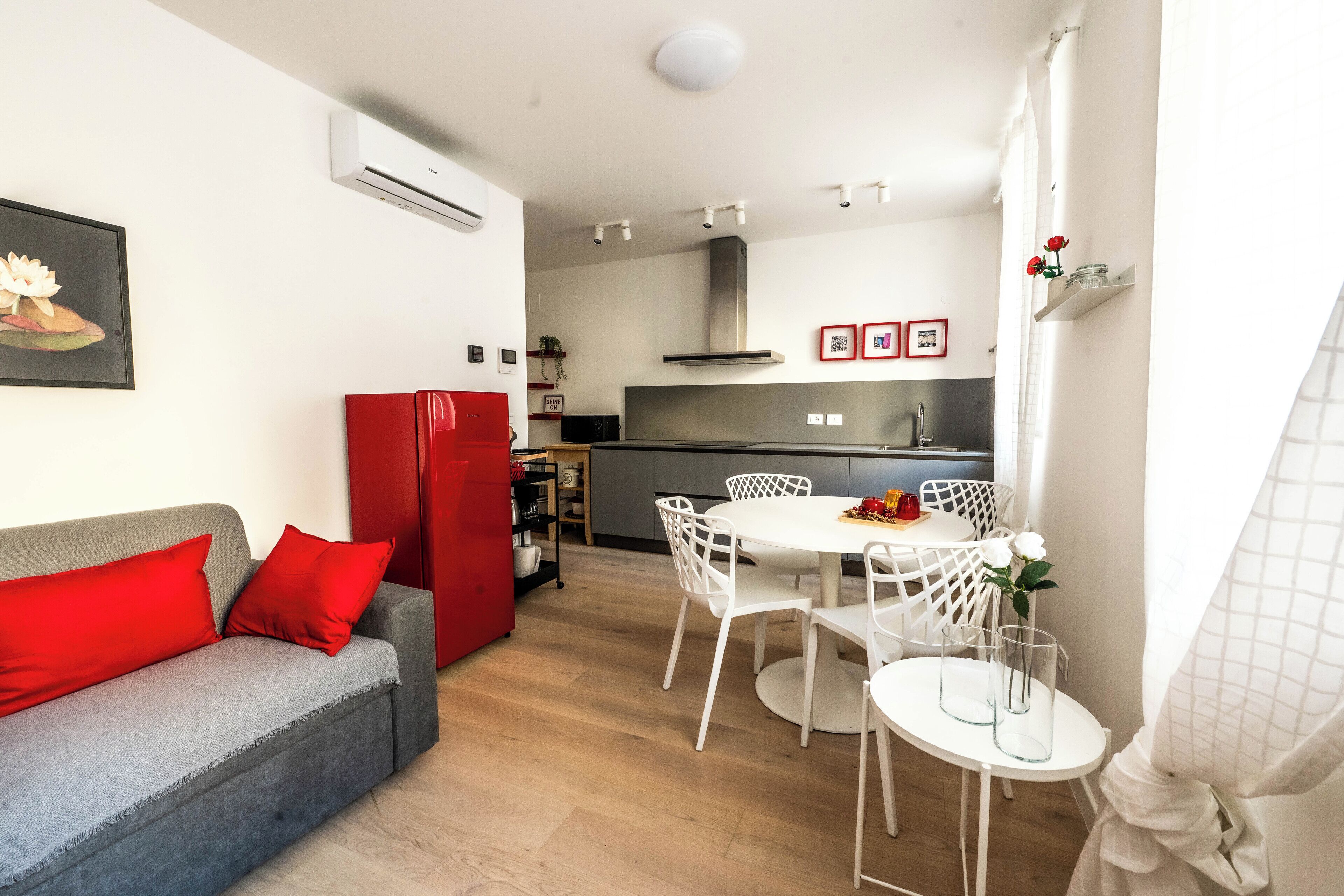 Un espacio habitable compacto con un sofá gris, cojines rojos, una mesa de comedor blanca con sillas, un refrigerador rojo y una zona de cocina al fondo.