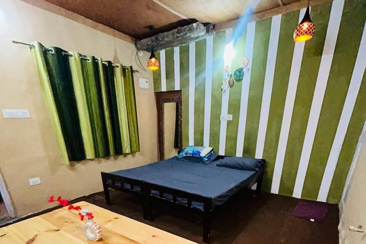 A small, cozy room with a bed, a wooden table, and a patterned stool.