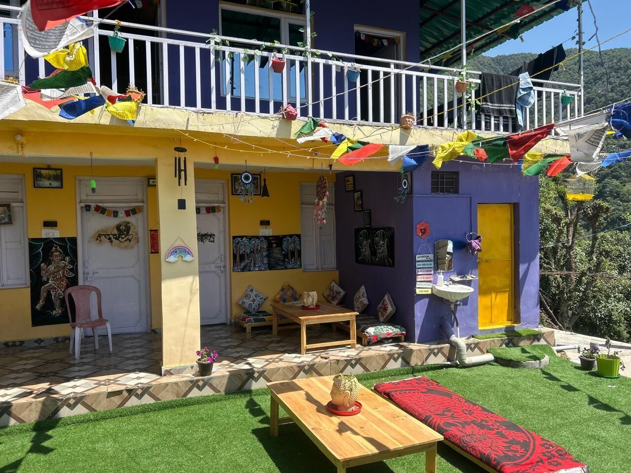 A colorful building with a balcony decorated with flags, a small garden area with artificial grass, and a wooden table with a red cushion.