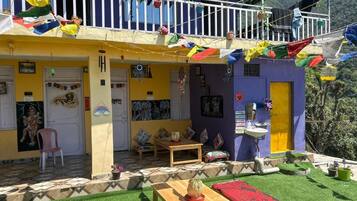 A colorful building with a balcony decorated with flags, a small garden area with artificial grass, and a wooden table with a red cushion.