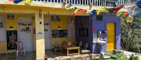 Un edificio colorido con un balcón adornado con banderas, un pequeño jardín con césped artificial y una mesa de madera con cojín rojo.
