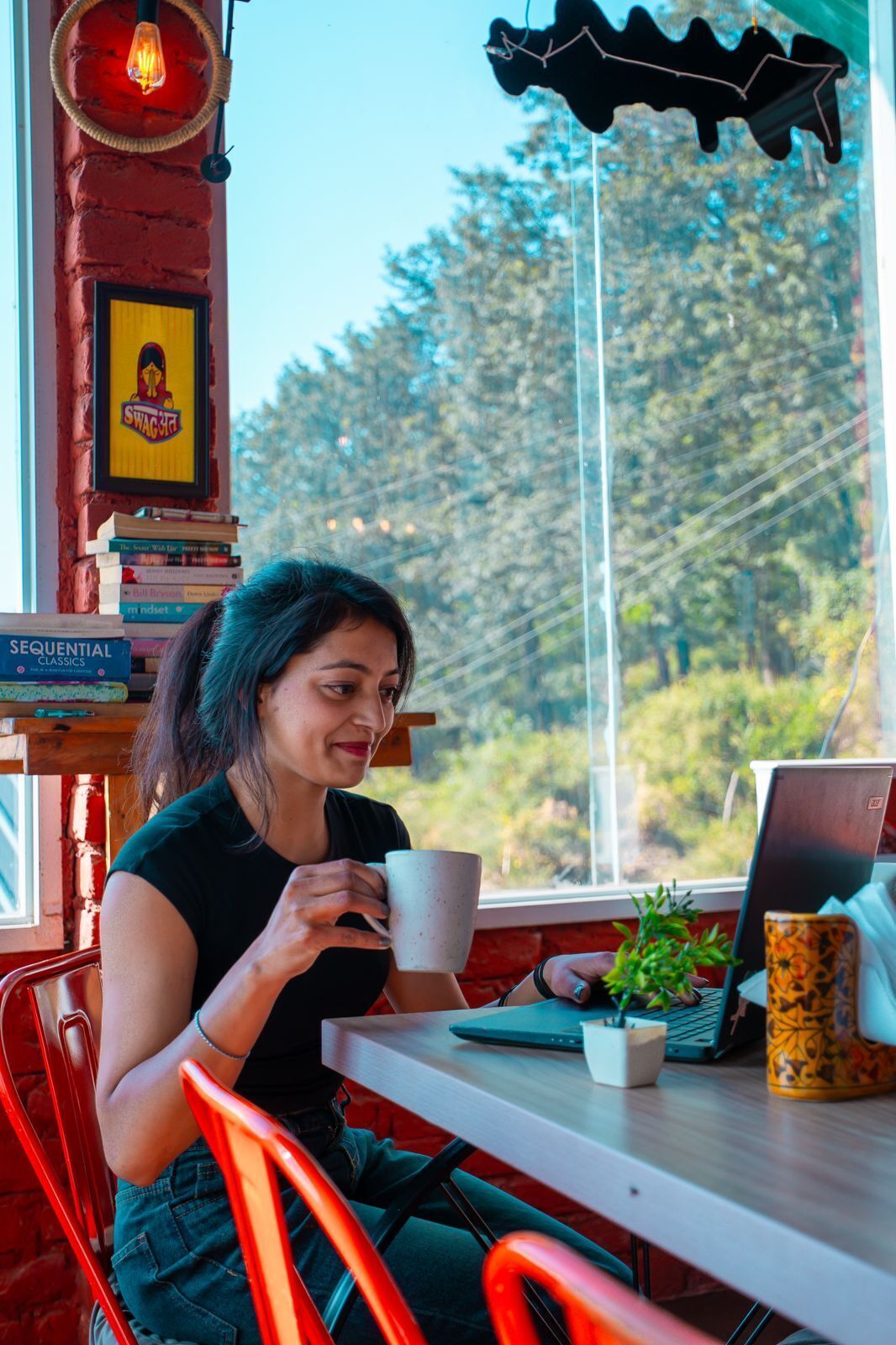 A person sitting at a table with a laptop, a mug, and a small plant. There is a bookshelf with books and a framed picture on the wall.