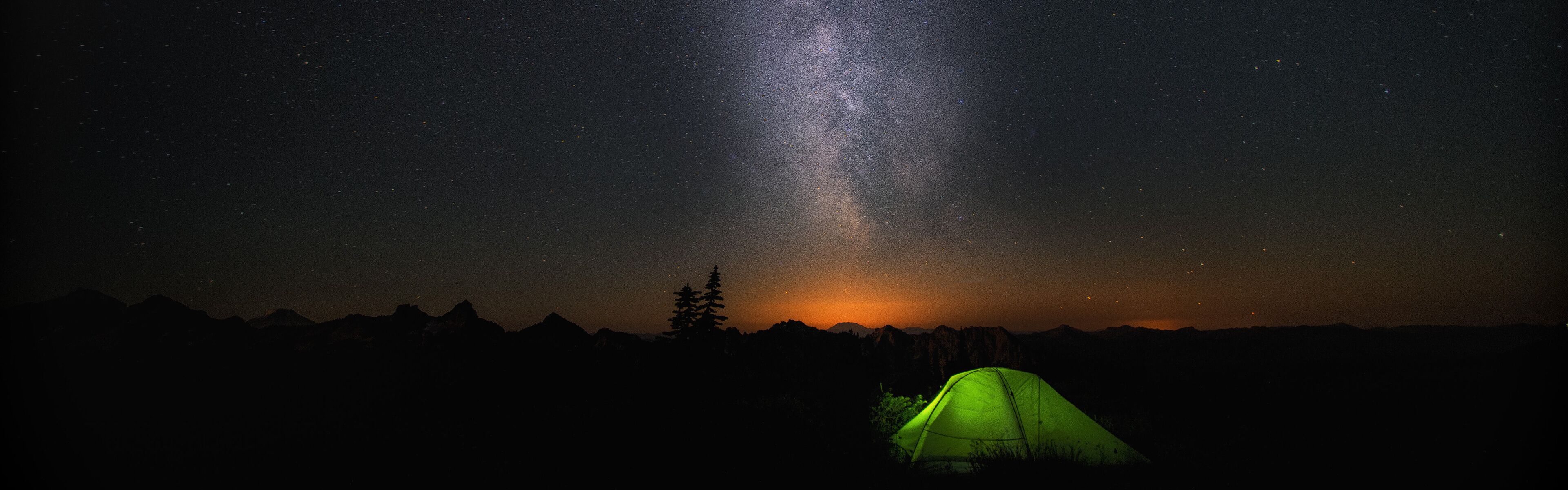 A night sky with stars and the Milky Way, a green tent, and a silhouette of mountains.