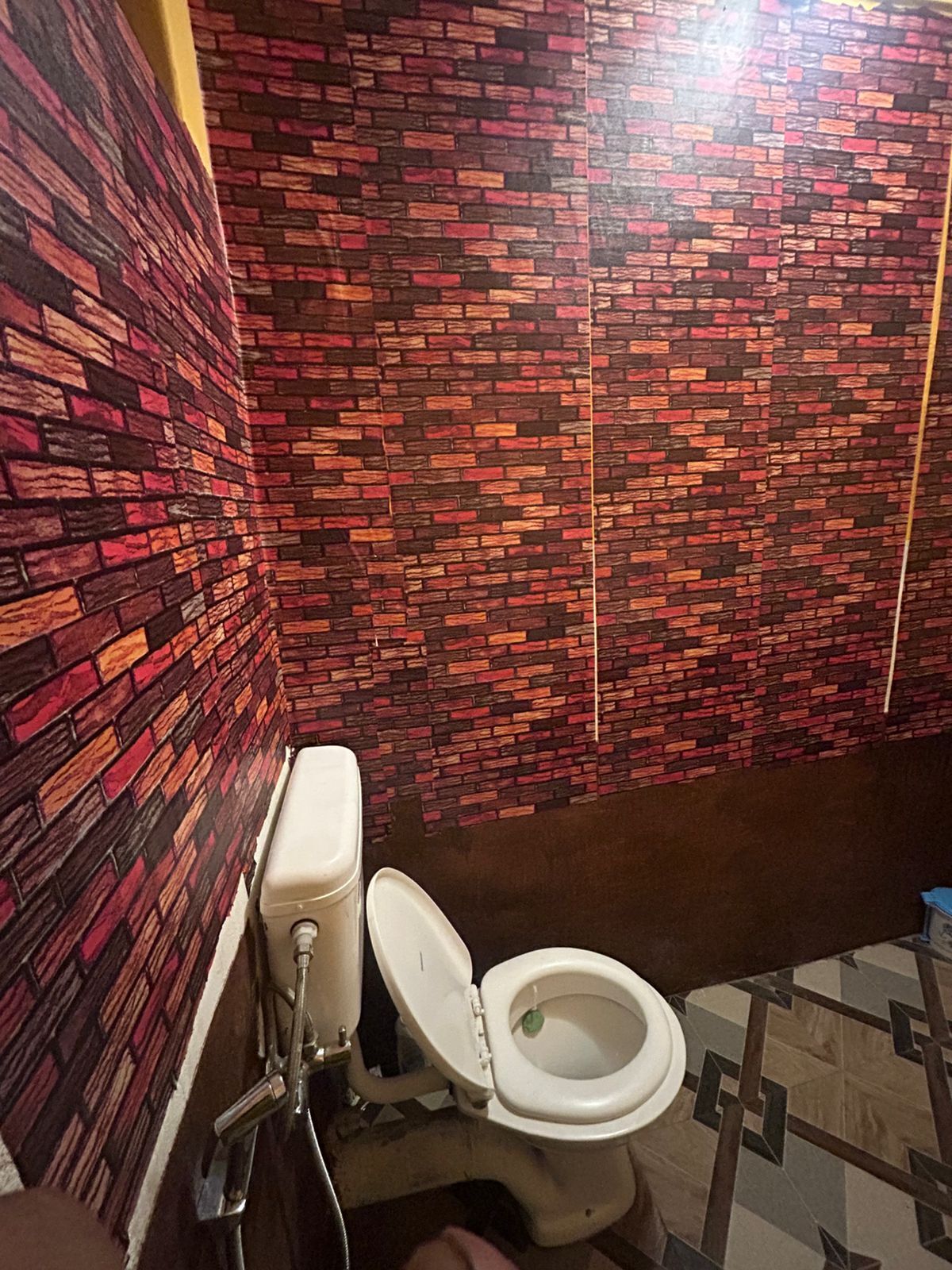 A bathroom with a toilet, a wall with a brick pattern, and a tiled floor.