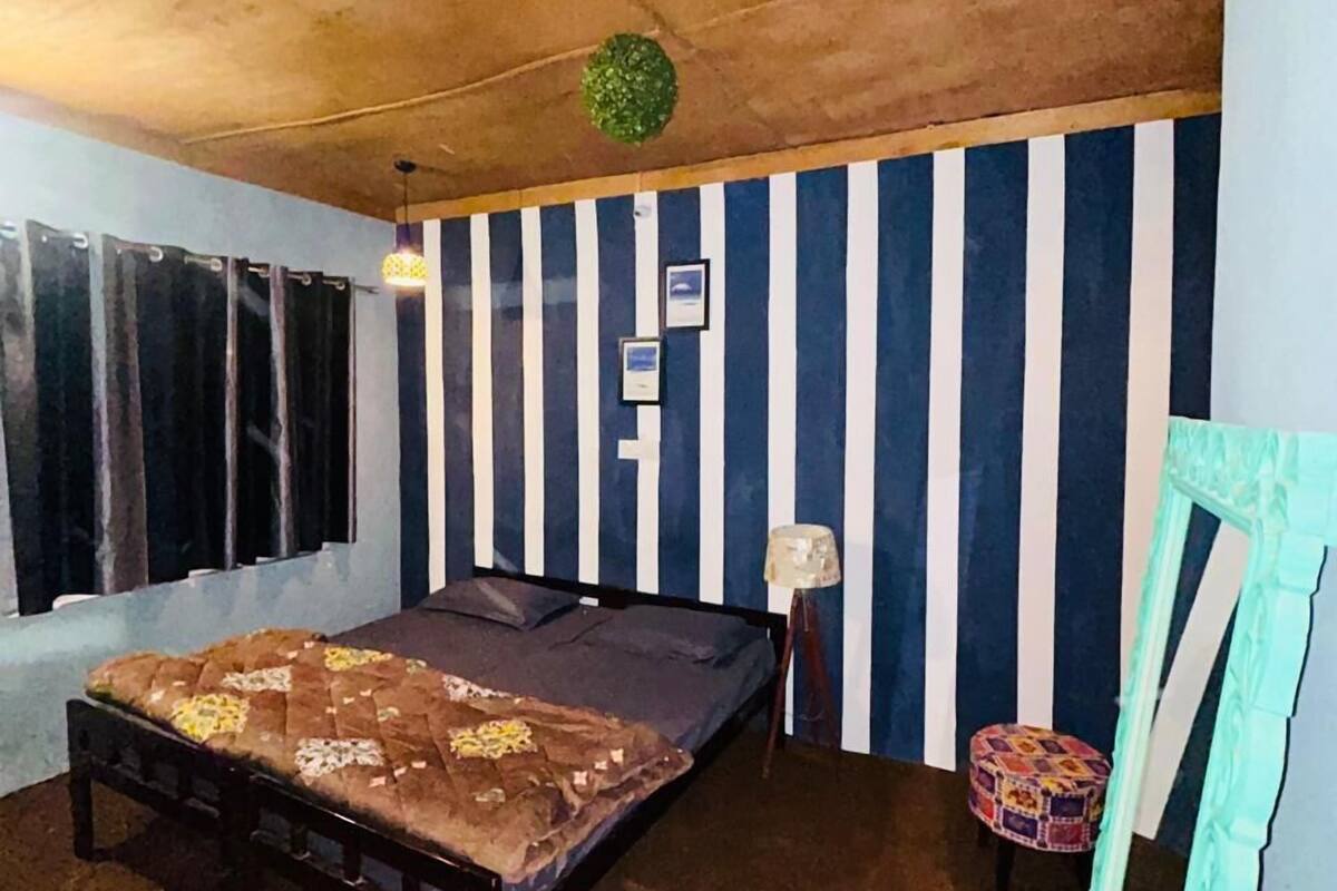 A room with a bed, a window with curtains, a striped wall, and a small stool.