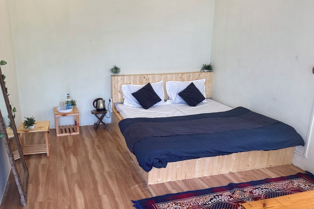 Une chambre à coucher avec un lit en bois, deux oreillers, une table de chevet avec une bouilloire, une petite plante et un élément décoratif mural.