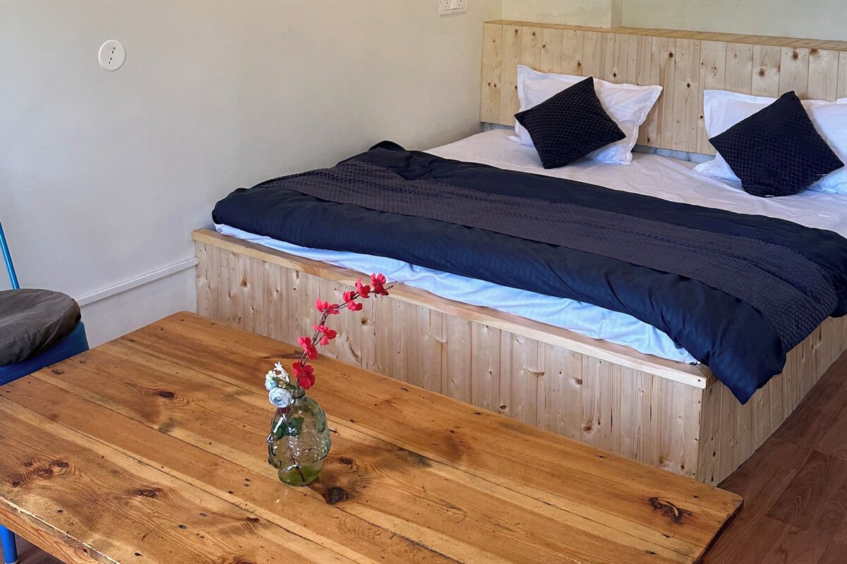 Une chambre à coucher avec un lit, une table en bois ornée d’un vase de fleurs et un ventilateur de plafond.