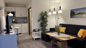En moderne stue med en sort sofa, et træsofabord og et hvidt sengebord. Der er et køkkenområde med hvide skabe og en mikrobølgeovn.