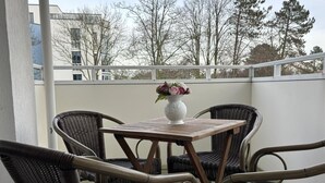En balkong med flätade stolar och ett bord, en vit vas med blommor och utsikt över byggnader och träd.
