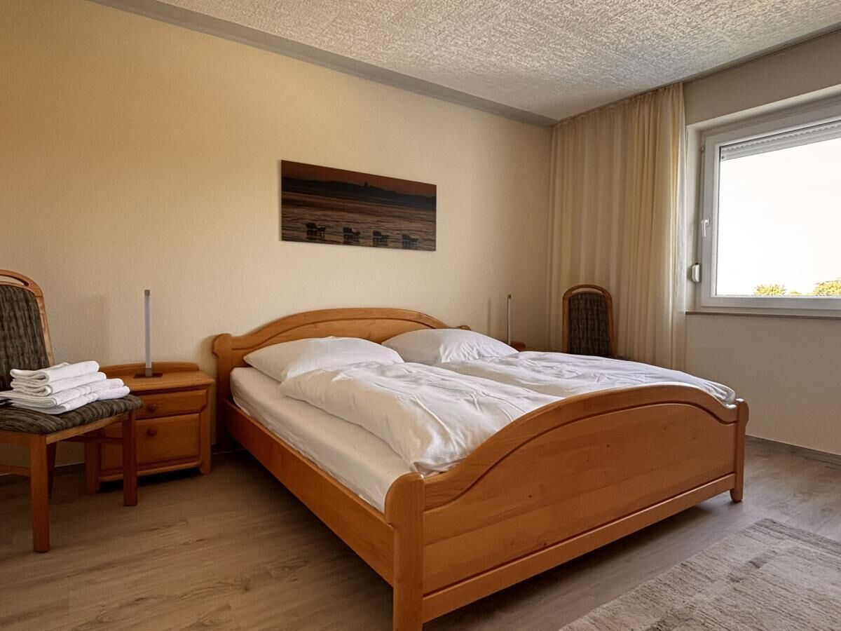 A bedroom with a wooden bed, a nightstand with a lamp, a chair, and a window with curtains.