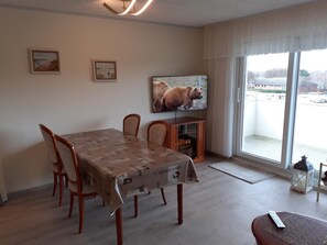 Ein Essbereich mit Tisch, der mit einer Tischdecke bedeckt ist, Holzstühlen, einem an der Wand befestigten Fernseher und Blick durch das Fenster.