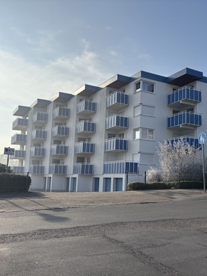 Ein mehrgeschossiges Wohngebäude mit Balkonen und einem Parkplatz davor, bewölkter Himmel.