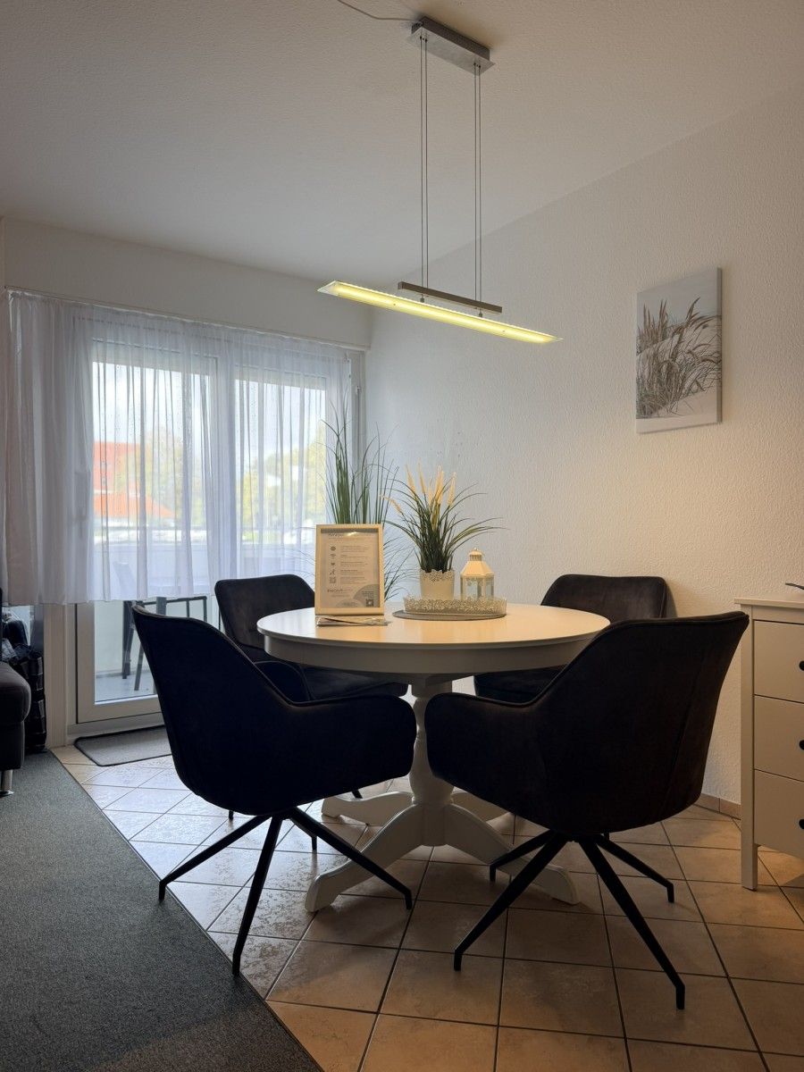 Uma área de refeições com uma mesa redonda, cadeiras pretas, um lustre e uma janela com cortinas translúcidas.
