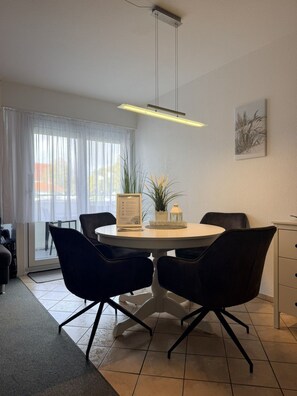 A dining area with a round table, black chairs, a hanging light fixture, and a window with sheer curtains. - Poseidon House, Apartment POS03 (Cuxhaven)