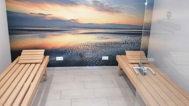 Une salle de spa avec une grande fresque représentant un coucher de soleil sur l’eau, deux bancs en bois et un sol carrelé.