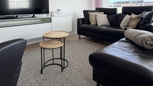 A modern living room with a black leather sofa, a small round table, a flat-screen TV mounted on the wall, and a window with a view of buildings. - House Poseidon, vacation apartment POS20 incl. beach chair (Cuxhaven)