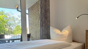 Ein Einzelbett mit Holzrahmen, einer weißen Matratze und einem weißen Kissen. Ein Fenster bietet Blick auf Bäume und ein Gebäude.